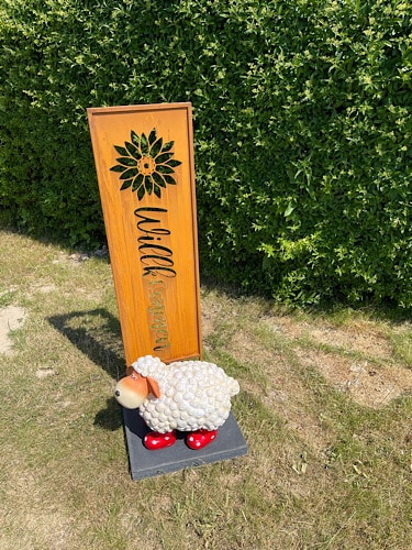 Schild mit Willkommen und ein Schaf aus Kunststoff davor mit kleinen roten Stiefeln